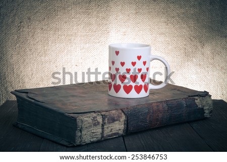 White mug with many pictured hearts and old book on the old wooden table on burlap background. Toned.
