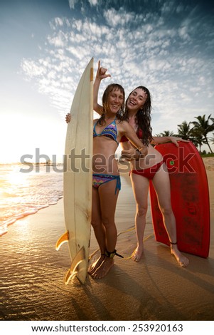 Two young ladies surfers standing with boards on the beach and showing shaka signs
