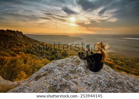 A woman on the top of a rock taking a picture using digital tablet