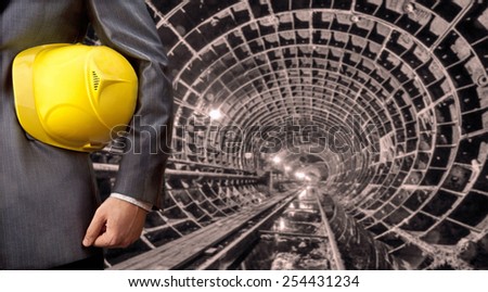 worker or engineer hand holding yellow helmet for workers security against the background of an underground mine with arc legs and rails for trolleys with coal in perspective