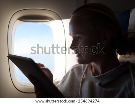 Woman typing on tablet computer sitting by illuminator in plane. Filling in time during the flight