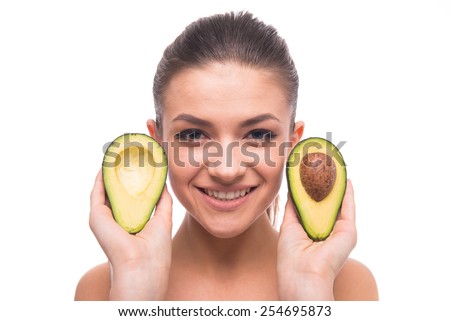 Young, smiling woman is holding avocado and looking at the camera on white background.