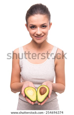 Young, smiling woman is holding avocado and looking at the camera on white background.