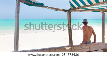 Man hiding in shade from burning tropical sun on picture perfect tropical sandy beach. Summer leisure. Shot from back.