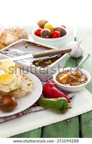 White pate dish with knife, bread, butter, chutney, tomatoes, peppers and garlic on red checkered cloth over green painted grunge style wooden table
