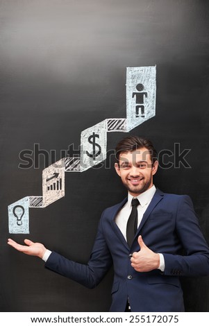 Handsome smiling businessman near dark chalkboard pointing on colorful symbols painted on it and showing thumb up