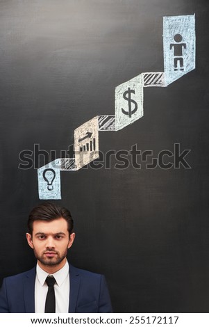 Serious young businessman standing near dark chalkboard with colorful symbols painted on it