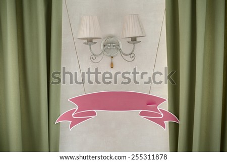 Blank pink wedding plate in the form of ribbons hanging on the wall by wall lamp. Front view.