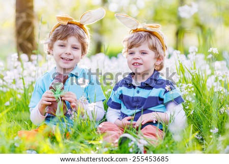 Family of two siblings: Little kid boys wearing Easter bunny ears at spring green grass and blooming apple garden, eating chocolate bunny and having fun outdoors. Celebrating traditional holiday.