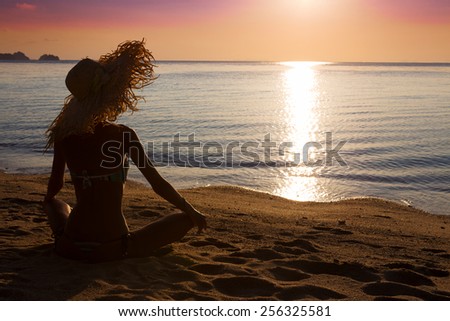 Slim woman in big hat is sitting on the beach and looking to the sunset