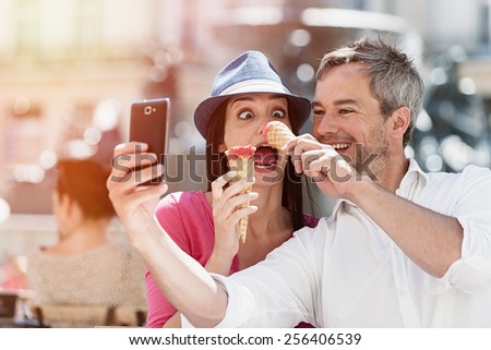 Portrait of a smiling couple eating ice cream and having fun in the city. The grey hair man with a beard is taking a selfie with phone. they make funny faces