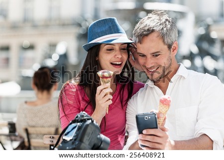 Portrait of a smiling couple eating ice cream in the city. The grey hair man with a beard in a white shirt is looking to his phone. The woman is wearing a blue hat and a pink top.