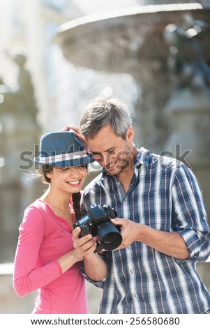 A smiling couple is looking at their photos on the screen of their camera in the city center. The woman is wearing a blue hat. The grey hair man with a beard is wearing a black backpack.