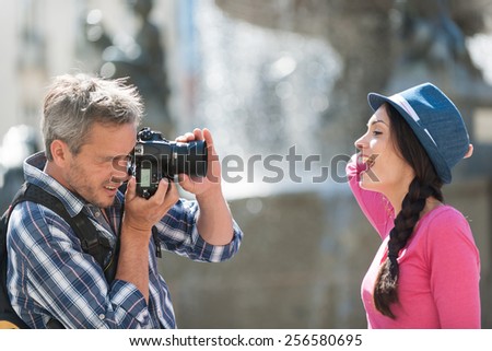 A loving couple is taking photos in the city center. The grey hair man with a beard and a black backpack is taking a closed picture of the smiling woman with a blue hat and a pink top.