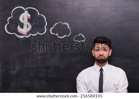 Photo of handsome young businessman on chalkboard background. Man sadly looking at camera. Cloud formed dialog and dollar sign in it painted on chalkboard