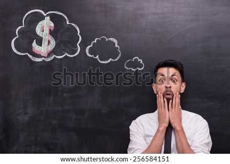 Photo of handsome young businessman on chalkboard background. Man anxiously looking at camera. Cloud formed dialog and dollar sign in it painted on chalkboard