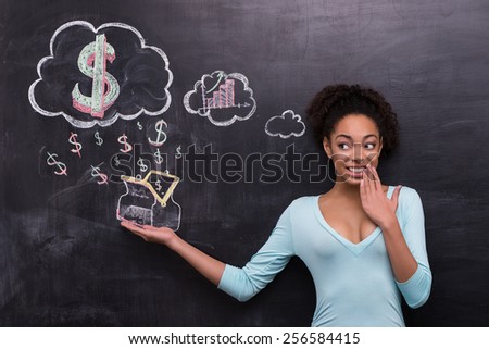 Photo of young afro-american woman on chalkboard background. Woman smiling and cheerfully looking at dollar signs and purse painted on chalkboard