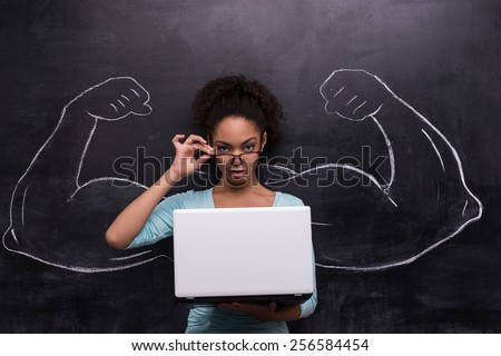 Funny picture of young afro-american woman with laptop on chalkboard background. Two strong muscular arms painted on chalkboard