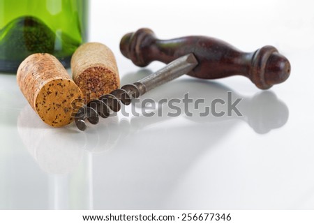 Close up front view of antique corkscrew, focus on front part of corkscrew and cork on far left side, on glass table with green empty wine bottle in background along with reflections 