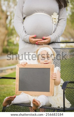 Pregnant Mom Behind Cute Baby Girl Sitting in Chair Holding Blank Blackboard.