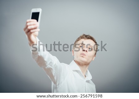 Handsome young man in shirt holding camera and making selfie and smiling while standing against grey background