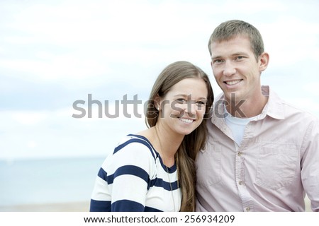 A beautiful young happy caucasian couple posing with the beach in the background on a cloudy day.