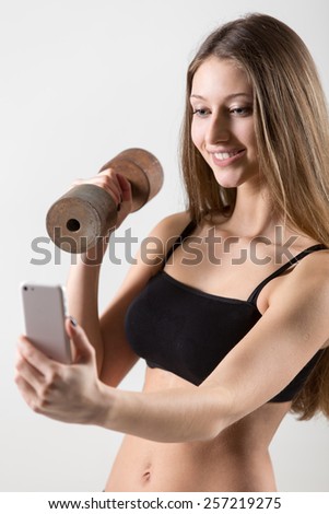 Smiling sporty teenage girl doing dumbbell curls and using app on mobile phone, taking picture, selfie, self-portrait with smartphone, reading text