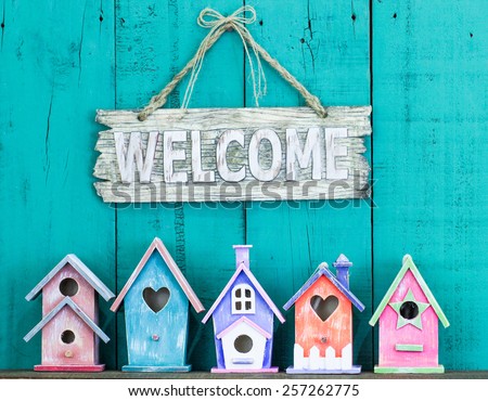 Wood welcome sign hanging over row of colorful spring birdhouses with antique teal blue rustic wooden background; purple, orange, pink, green 