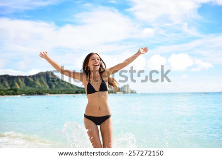 Beach fun woman happy on Hawaii vacations. Cheering Asian girl in black bikini with arms up in happiness playing and running in water on Waikiki beach, Honolulu city, Oahu island, Hawaii, USA.