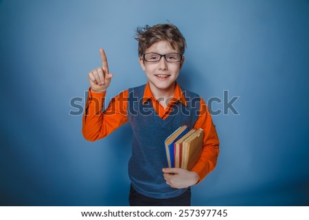 boy teenager with books of European appearance with glasses raised his finger smiling on a gray background, retro, joy