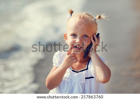 A portrait of a cute little smiling blonde girl in a white dress calling by phone standing on the beach on a warm summer day. Holiday on a seashore concept. Funny kids. Sea. Ocean.
