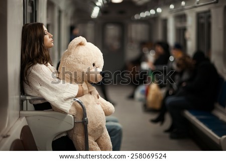 Sad girl hugged her toy bear and riding in a subway car. The background blured, people are not recognizable. Photographs of the executed on the open aperture with a soft focus. Photo toned. Royalty-Free Stock Photo #258069524