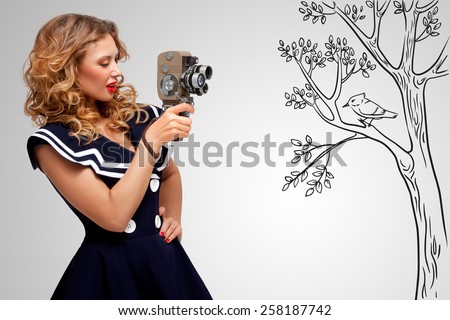 Glamorous pin-up sailor girl filming nature and wildlife with an old retro cinema 8 mm camera, standing in front of a bird on grey sketchy background. Royalty-Free Stock Photo #258187742