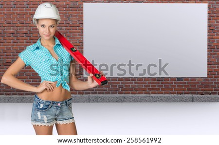 Woman in helmet holding spirit level, smiling. Brick wall and empty board as backdrop