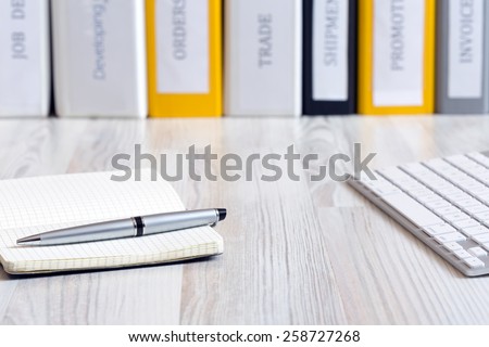Desk of decision maker.
Side view on light wooded desk with pen, stylish leaver note pad and keyboard, with stack of folders on the background