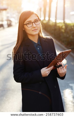 Charming female student standing outdoors with her digital tablet pc, stylish young woman standing on beautiful city road at sunny spring evening and holding gadget in the hands