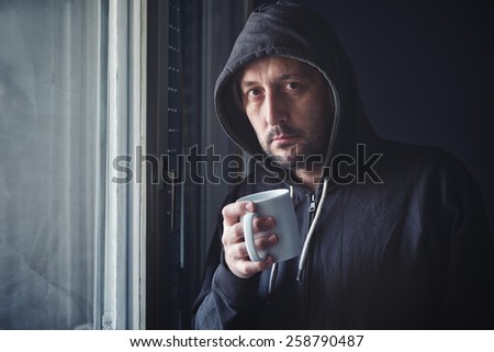 Adult Man with Hooded Jacket Holding Cup of Hot Coffee by the Window, Drinking Beverage and Making Plans for the Day Ahead. Selective Focus with Shallow Depth of Field.