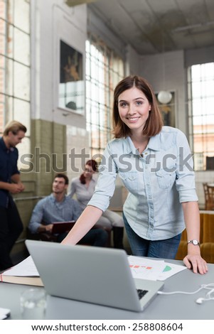 Smiling woman working in a design studio standing at her desk with a laptop and large paper drawing while her colleagues work in the background Royalty-Free Stock Photo #258808604