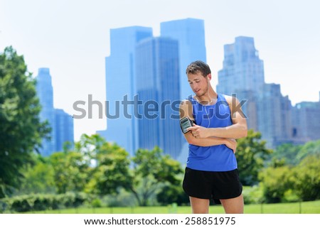 New York City man runner listening music on smartphone. Male adult jogger running using touchscreen on armband for workout in Central Park with urban background of Manhattan's skyscrapers skyline.