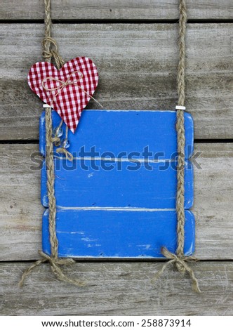 Blank antique blue sign with red gingham heart hanging by braided rope on rustic wooden background