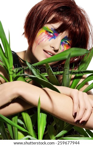 Fashion model with short dark fluffy hair and colorful make-up posing with a green plant.