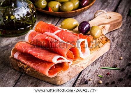 thin slices of prosciutto with mixed olives on wooden cutting board Royalty-Free Stock Photo #260425739