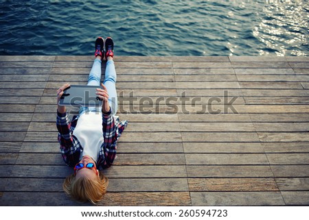 Young teen woman relaxing upon a wooden jetty while taking self portrait holding up tablet, female tourist making remembering portrait during her holidays, surrealistic photo, filter, cross process