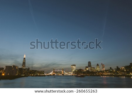 London skyline at dusk with River Thames on foreground. From left to right The Shard, Tower Bridge, Walkie Talkie and other skyscrapers. All buildings have lights on.