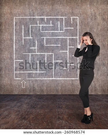 Young businesswoman looking at the labyrinth and thinking about right way on grey brick background