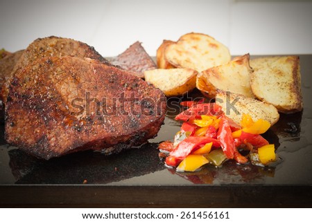 Juicy grilled steaks served with roasted potatoes and peppers on black granite board, vignetted
