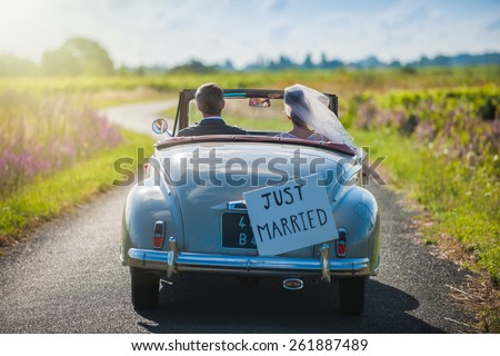 A newlywed couple is driving a convertible retro car on a country road for their honeymoon, rear view Royalty-Free Stock Photo #261887489