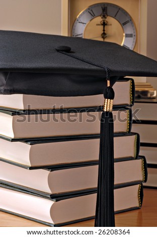 Graduation cap on stack of books with clock in background.  Softly lit still life with shallow dof.