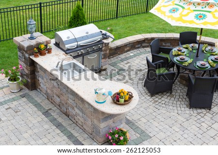 High angle view of a stylish outdoor kitchen, gas barbecue and dining table set for entertaining guests with formal place settings and flowers on a paved patio