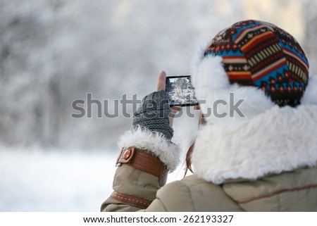 Women in winter clothing, fingerless mittens and ornamented hat photographing snowy forest by a mobile phone. Shallow dof. Focus on hand.